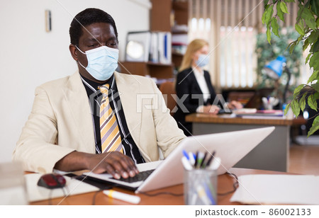 Businessman in protective mask working alone with laptop and papers 86002133