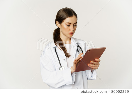 Woman doctor looking concerned at digital tablet, reading with worried face expression, wearing white coat, standing over white background 86002259
