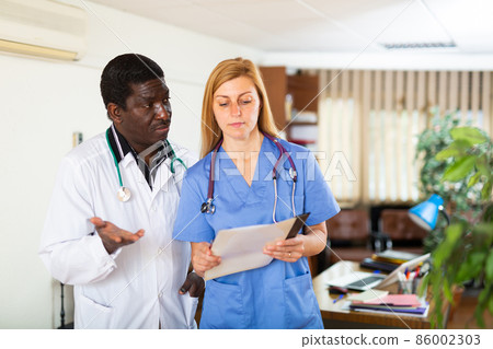 Two multinational doctors in the clinic discuss work issues while standing in an office 86002303