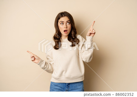 Confused girl shopper pointing sideways, making choice, decision between two variants, beige background 86002352