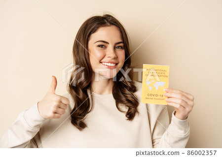 Covid-19 vaccination concept. Portrait of happy healthy girl showing coronavirus international vaccination certificate and thumb up, travelling during pandemic, beige background 86002357