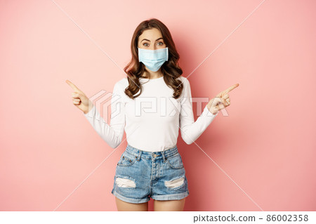 Portrait of beautiful woman in face medical mask pointing sideways, showing two choices, ways, sides, demonstrating advertisement, pink background 86002358