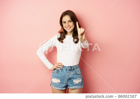 Only one. Smiling beautiful and stylish woman showing index finger, looking confident, prohibit stop something, standing happy against pink background 86002387