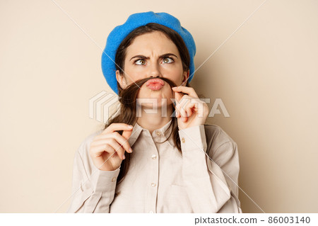 Funny beautiful girl fooling around, making moustache out of hair under lips, squinting and grimacing, standing happy against beige background Funny beautiful girl fooling around, making moustache out of hair under lips, squinting and grimacing, standing happy against beige background 86003140