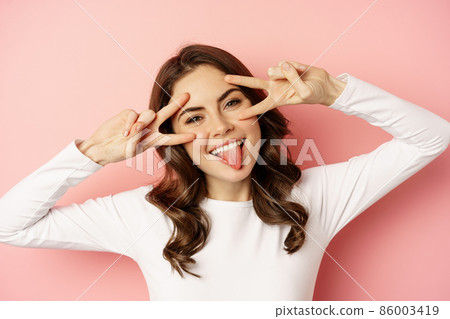 Close up face of young stylish girl, sassy woman showing happy, excited face expression, peace v-sign over eyes, standing against pink background 86003419