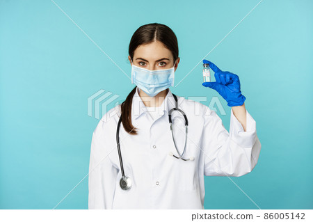 Vaccination campaign. Smiling woman doctor in face medical mask and gloves, showing vaccine from covid, omicron delta variant, standing over torquoise background 86005142