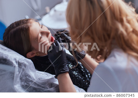 Caucasian young child girl sitting in medical chair while dentist fixing teeth at dental clinic using dental tools instruments, close-up photo of face. professional medical specialist. 86007345