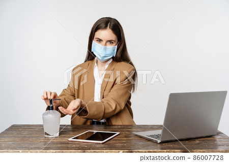 Businesswoman at workplace wearing face mask from covid-19, work in office during pandemic, using hand sanitizer to clean hands, posing over white background 86007728