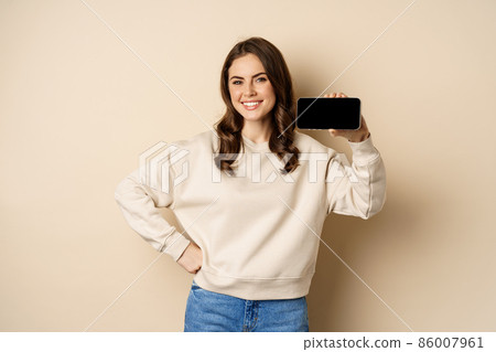 Portrait of smiling cute woman showing horizontal smartphone screen, website or application, standing over beige background Portrait of smiling cute woman showing horizontal smartphone screen, website or application, standing over beige background 86007961