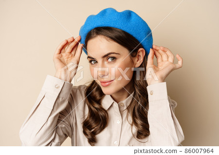 Stylish modern girl put on trendy hat on head and smiling, going out, posing against beige background 86007964