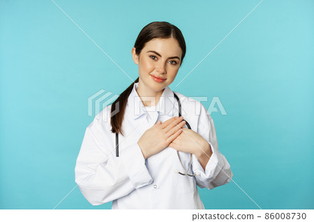 Caring healthcare worker, young female doctor holding hands on heart and smiling, looking with care and tenderness, standing over torquoise background Caring healthcare worker, young female doctor holding hands on heart and smiling, looking with care and tenderness, standing over torquoise background 86008730