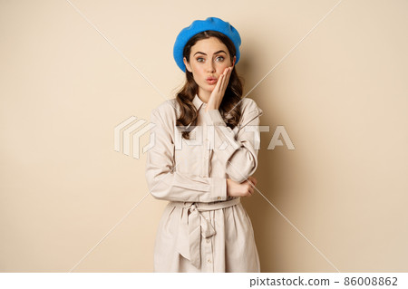Image of stylsh beautiful woman looking surprised, shocked reaction at camera, posing in trench coat against beige background 86008862