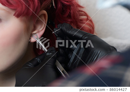 A beautician makes a puncture of the earlobe with a disposable device in a beauty salon. Ear piercing lobe. 86014427