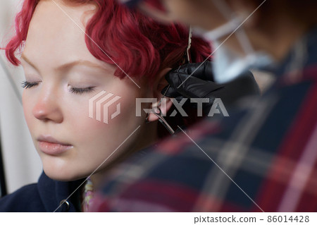 A beautician makes a puncture of the earlobe with a disposable device in a beauty salon. Ear piercing lobe. A beautician makes a puncture of the earlobe with a disposable device in a beauty salon. Ear piercing lobe. 86014428