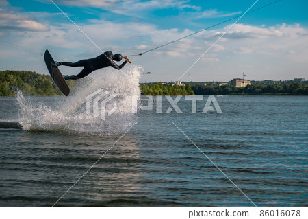 Wakeboarder practicing tricks jumping on board above water surface creating splashes Wakeboarder practicing tricks jumping on board above water surface creating splashes 86016078