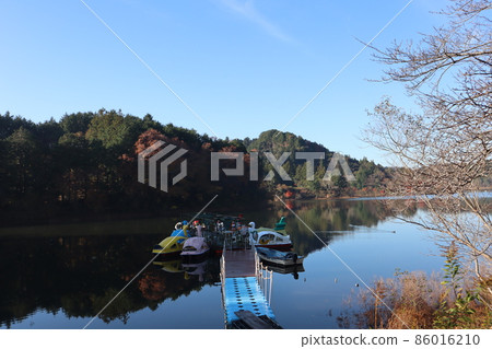 捕捉天鵝船登陸、藍天、薄雲、秋葉在冬季枯樹和枯草前的山景的池塘景色。 捕捉天鵝船登陸、藍天、薄雲、秋葉在冬季枯樹和枯草前的山景的池塘景色。 86016210