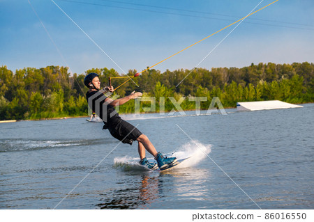Focused man sliding on wakeboard on water surface holding tow rope with one hand Focused man sliding on wakeboard on water surface holding tow rope with one hand 86016550