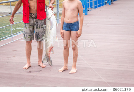 Fisherman holding a huge grass carp on a pier on a lake, standing with his son Fisherman holding a huge grass carp on a pier on a lake, standing with his son 86021379