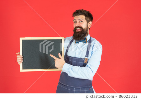 Happy guy in barber apron smile pointing finger at blackboard red background copy space, barbershop Happy guy in barber apron smile pointing finger at blackboard red background copy space, barbershop 86023312