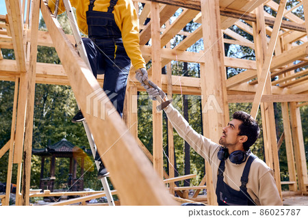 bearded male in uniform helps female constructor standing in height, man giving hammer bearded male in uniform helps female constructor standing in height, man giving hammer 86025787