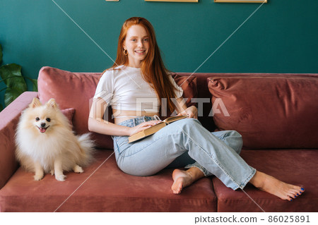 Portrait of smiling young woman reading book sitting on cozy sofa with pretty spitz pet dog at apartment with modern interior, looking at camera. Happy female spending pleasant weekend with doggy. Portrait of smiling young woman reading book sitting on cozy sofa with pretty spitz pet dog at apartment with modern interior, looking at camera. Happy female spending pleasant weekend with doggy. 86025891
