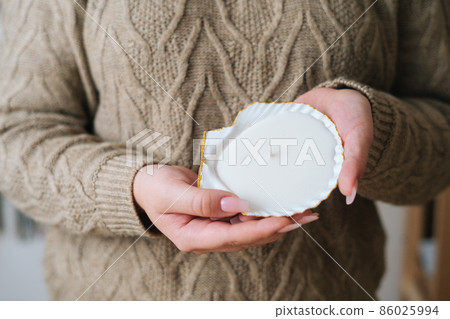 Close-up cropped shot of unrecognizable young woman holding scented handmade candle in candlestick made of exotic seashell. Female wearing sweater holding aroma relaxing candle in hand. Close-up cropped shot of unrecognizable young woman holding scented handmade candle in candlestick made of exotic seashell. Female wearing sweater holding aroma relaxing candle in hand. 86025994