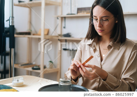 Close-up portrait of focused female artisan using piece of beeswax to create wick for handmade candle at workshop. Process of making hand-made natural candle at home. Close-up portrait of focused female artisan using piece of beeswax to create wick for handmade candle at workshop. Process of making hand-made natural candle at home. 86025997