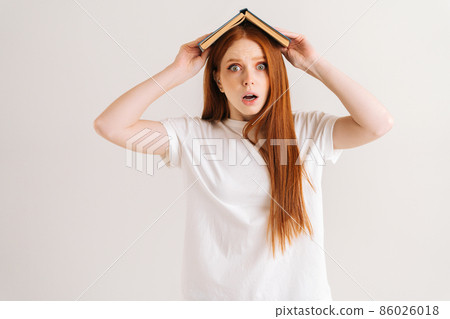 Studio portrait of afraid young student woman covering head with book looking at camera, standing on white isolated background. Shocked redhead lady posing with educational materials. Studio portrait of afraid young student woman covering head with book looking at camera, standing on white isolated background. Shocked redhead lady posing with educational materials. 86026018