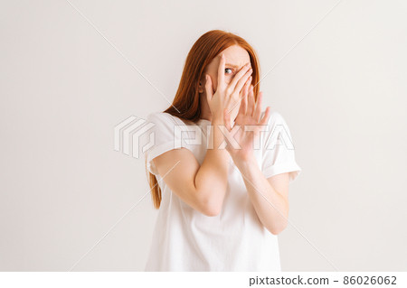 Studio portrait of frightened redhead young woman looking shy or terrified covering hiding face with hands peeping through fingers, shocked by horror movie or frightened on white isolated background. 86026062