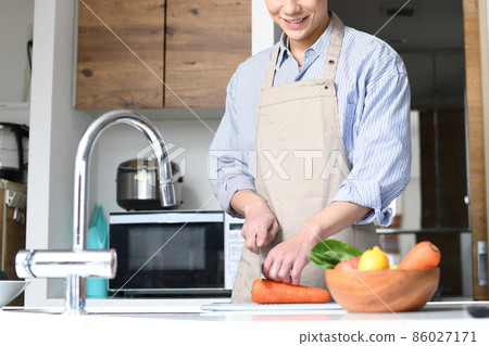 A young black-haired man in an apron cooking in the kitchen 86027171