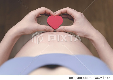 A pregnant woman after IVF holds a small red heart in her hands. The concept of motherhood, fertility, new life, In vitro fertilisation. 86031852