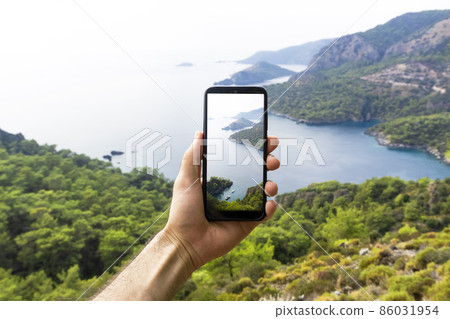 A hand with a smartphone taking pictures of the natural landscape, sea and mountains on a journey. Phone, internet and mobile network concept. Impressions and Adventures. A hand with a smartphone taking pictures of the natural landscape, sea and mountains on a journey. Phone, internet and mobile network concept. Impressions and Adventures. 86031954
