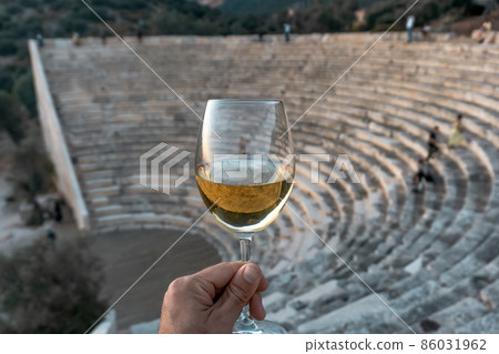 A glass of white wine in a man's hand. Greek Mediterranean wine against the backdrop of an antique amphitheater in Greece. 86031962