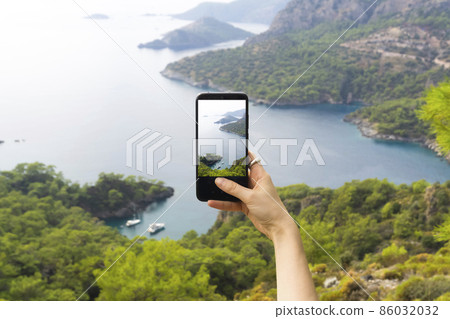 A hand with a smartphone taking pictures of the natural landscape, sea and mountains on a journey. Phone, internet and mobile network concept. Impressions and Adventures. 86032032