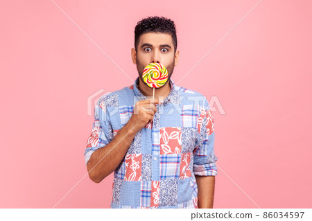 Young adult bearded man wearing blue casual shirt covering his astonished face with round sweet sugary rainbow candy, looking at camera with big eyes. Indoor studio shot isolated on pink background. Young adult bearded man wearing blue casual shirt covering his astonished face with round sweet sugary rainbow candy, looking at camera with big eyes. Indoor studio shot isolated on pink background. 86034597