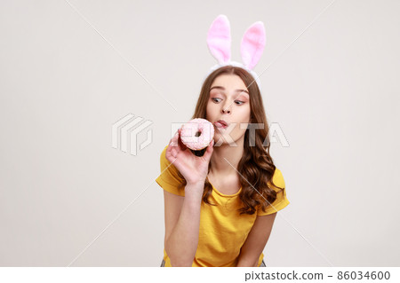 Portrait of funny young woman with brown hair playfully showing tongue out to lick donut, having pink bunny ears, humorous expression. Indoor studio shot isolated on gray background. 86034600