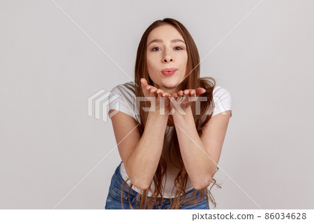 Catch my kiss. Lovely romantic woman looking at camera, sending air kissing over palms hands, expressing love, wearing white T-shirt. Indoor studio shot isolated on gray background. 86034628
