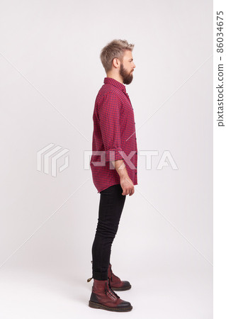Side view full body length of serious confident bearded male wearing red plaid shirt and casual black pants posing, practicing modeling. Indoor studio shot isolated on gray background 86034675