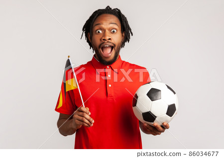 Amazed man with dreadlocks wearing red casual style T-shirt, supporting german soccer team on championship, cheering and greeting, patriotism. Indoor studio shot isolated on gray background. 86034677
