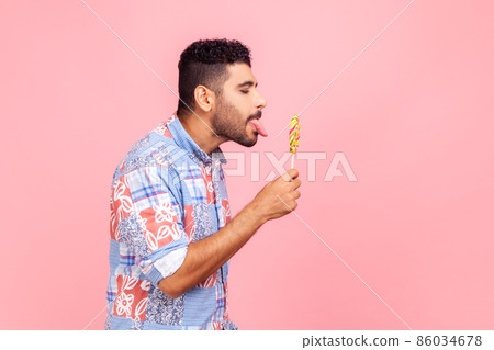 Side view of bearded handsome man wearing blue casual shirt standing tastes lollipop, dreaming licking round sweet sugary rainbow candy. Indoor studio shot isolated on pink background. 86034678