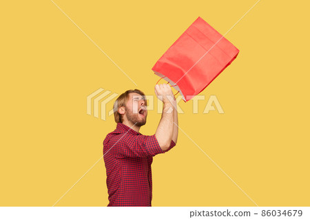 Portrait of unhappy hipster bearded guy wearing checkered shirt standing with empty shopping bag, looking inside trying to find at least something. Indoor studio shot isolated on yellow background. 86034679