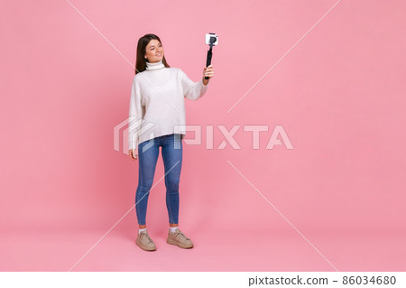 Full length portrait of happy young adult woman streaming, talking with followers, using steadicam, wearing white casual style sweater. Indoor studio shot isolated on pink background. Full length portrait of happy young adult woman streaming, talking with followers, using steadicam, wearing white casual style sweater. Indoor studio shot isolated on pink background. 86034680