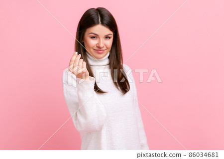 Attractive young adult woman standing and looking at camera with money or italian gesture with hand, wearing white casual style sweater. Indoor studio shot isolated on pink background. 86034681