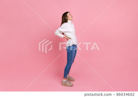 Full length portrait of girl touches injured back, suffers pain in lower lumbar muscular, arthritis, wearing white casual style sweater. Indoor studio shot isolated on pink background. 86034682