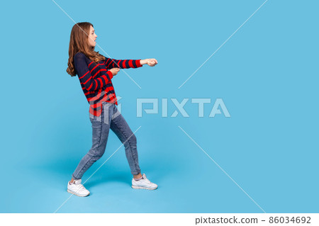Full length profile portrait of woman wearing striped casual style sweater, standing in attack or pulling hands gesture and looking forward. Indoor studio shot isolated on blue background. Full length profile portrait of woman wearing striped casual style sweater, standing in attack or pulling hands gesture and looking forward. Indoor studio shot isolated on blue background. 86034692