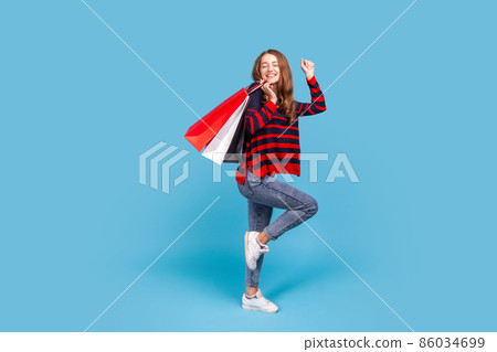 Full length portrait of positive woman wearing striped casual style sweater, standing with shopping bags, dancing, satisfied with her purchase. Indoor studio shot isolated on blue background. 86034699