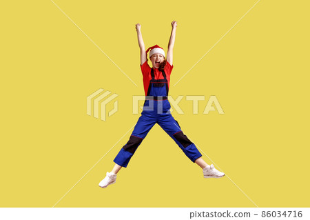 Full length portrait of excited happy female worker jumping high, finishing work before Christmas, wearing blue overalls and santa claus hat. Indoor studio shot isolated on yellow background. Full length portrait of excited happy female worker jumping high, finishing work before Christmas, wearing blue overalls and santa claus hat. Indoor studio shot isolated on yellow background. 86034716