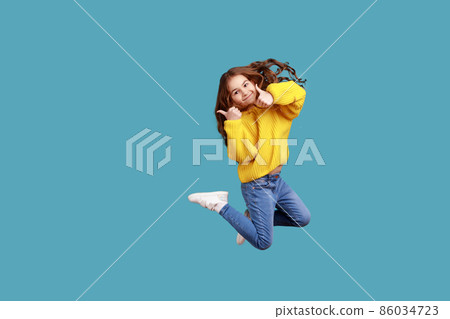 Full length portrait of happy charming little girl jumping up high and showing thumb up to camera, wearing yellow casual style sweater. Indoor studio shot isolated on blue background. Full length portrait of happy charming little girl jumping up high and showing thumb up to camera, wearing yellow casual style sweater. Indoor studio shot isolated on blue background. 86034723