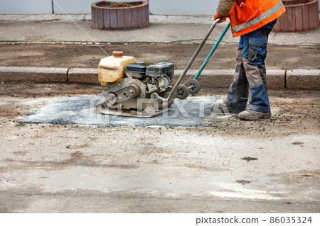 A worker with a petrol vibrating plate patches a pothole on an old road. 86035324