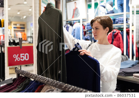 Young beautiful brunette woman with a short haircut in a white sweater chooses stylish clothes in a Young beautiful brunette woman with a short haircut in a white sweater chooses stylish clothes in a 86037531